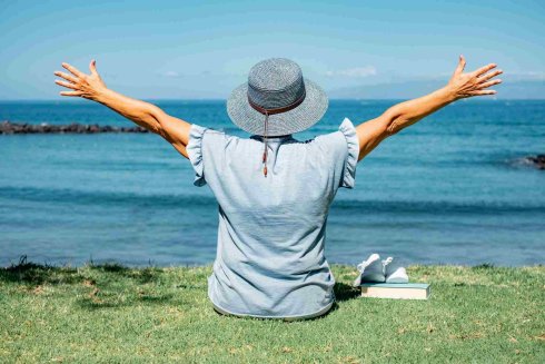 Back view of senior woman in hat sitting in meadow in front of sea looking at the horizon, relaxed senior lady enjoying free time on vacation or retirement Back view of senior woman in hat sitting in meadow in front of sea looking at the horizon, relaxed senior lady enjoying free time on vacation or retirement