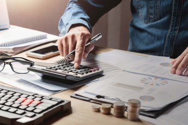 Buchhaltung Hand einer Person, die mit Taschenrechner und Unterlagen auf an einem Tisch arbeitet.