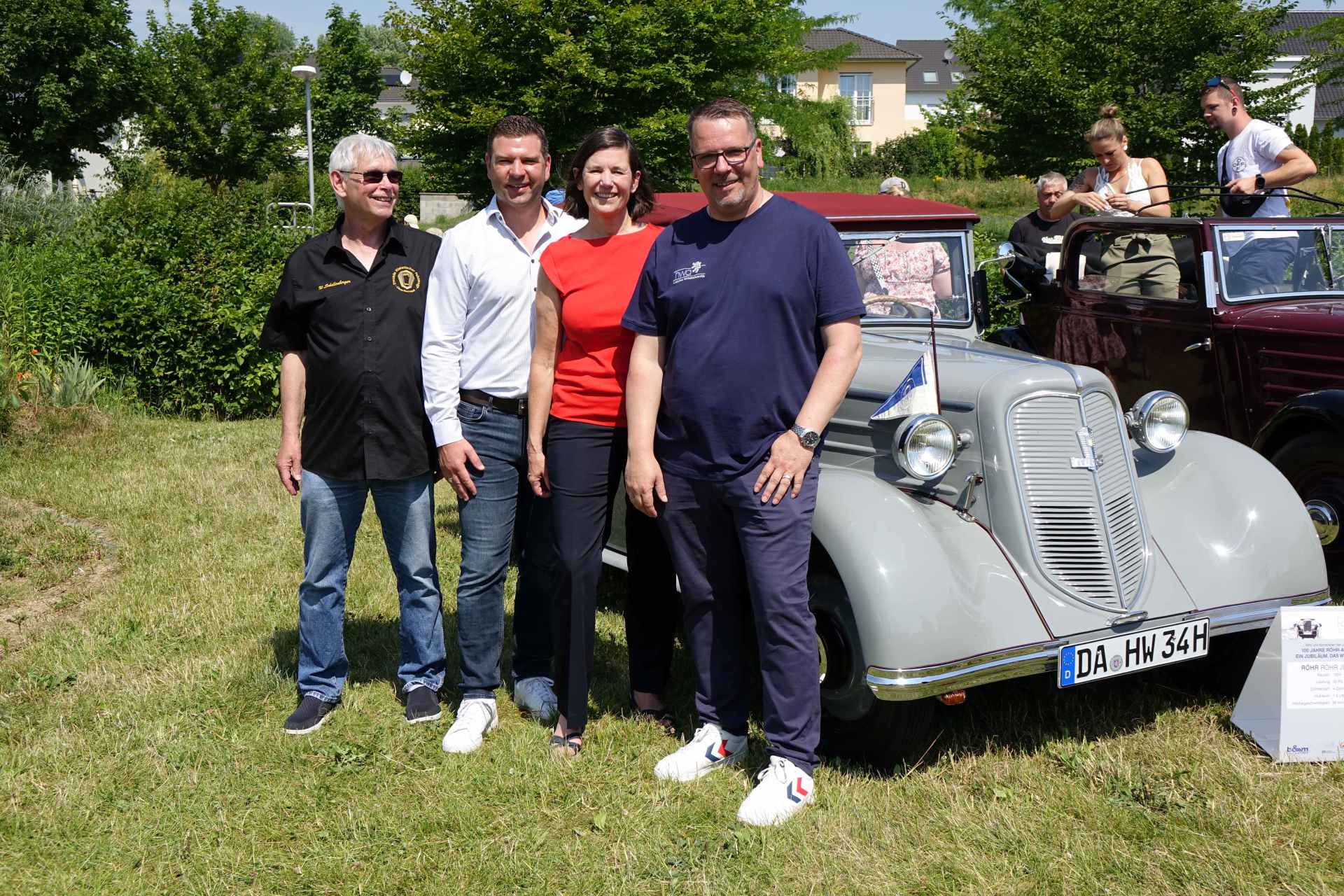 Gruppenfoto von Gästen und Organisatoren bei einer Jubiläumsveranstaltung mit historischem Fahrzeug im Freien.
