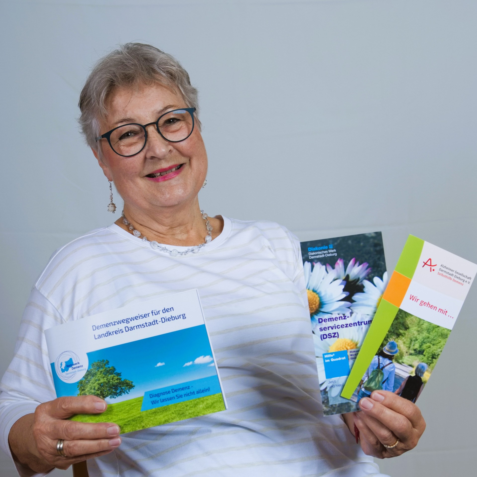 Lächelnde Frau hält Informationsbroschüren zu Demenz, Beratungsangeboten und Unterstützungsmöglichkeiten für Betroffene und Angehörige in den Händen.