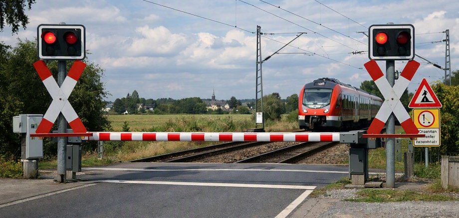„Geschlossener Bahnübergang mit Schranken und blinkenden Warnlichtern, Zug nähert sich im Hintergrund