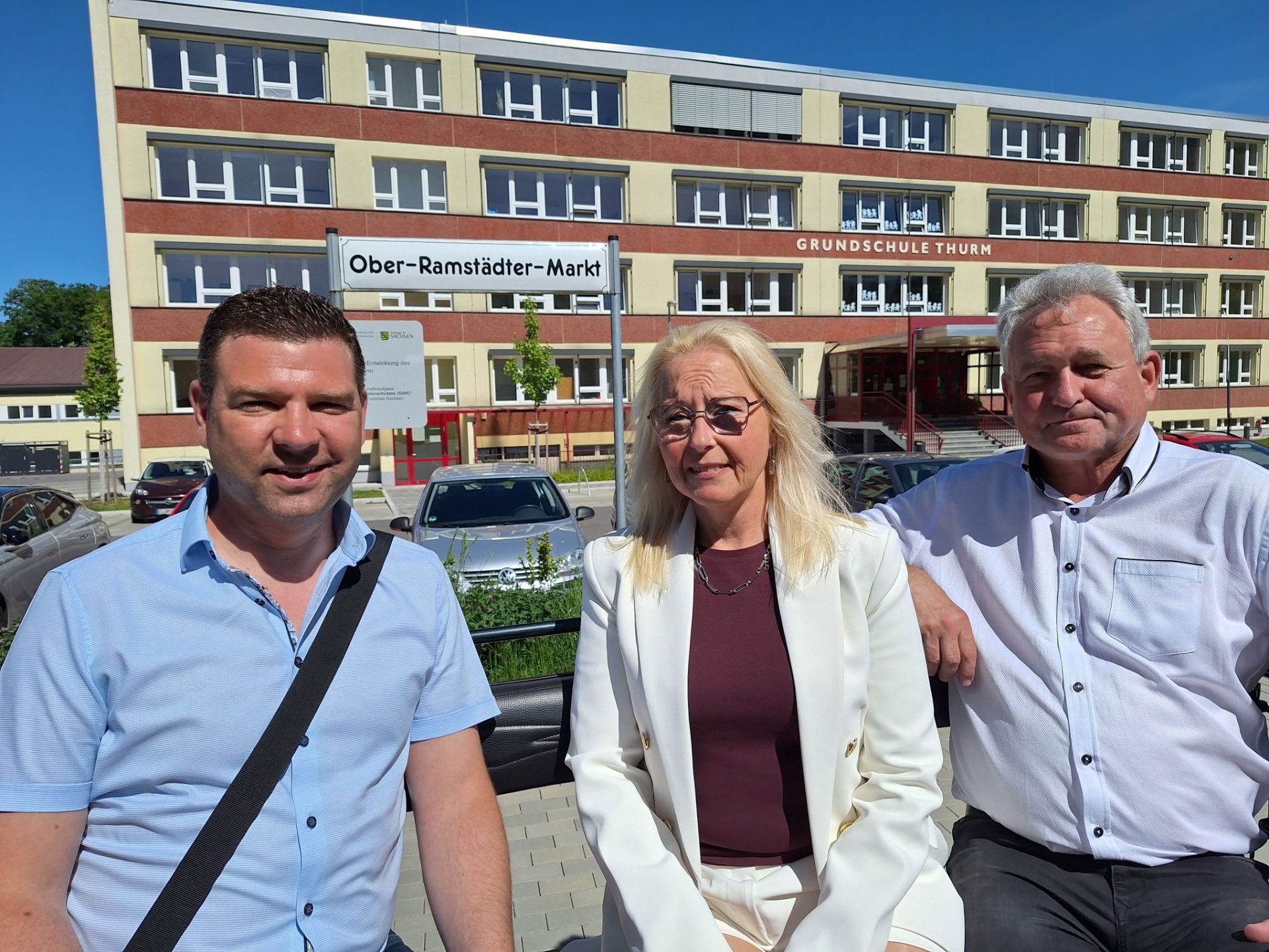 Drei Erwachsene sitzen vor einem mehrstöckigen Schulgebäude mit der Aufschrift ‚Grundschule Thurm‘, während im Vordergrund ein Straßenschild mit dem Namen Ober-Ramstädter-Markt zu sehen ist