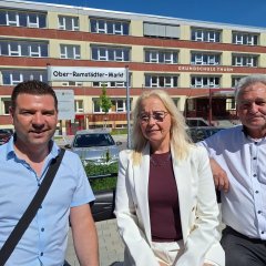 Drei Erwachsene sitzen vor einem mehrstöckigen Schulgebäude mit der Aufschrift ‚Grundschule Thurm‘, während im Vordergrund ein Straßenschild mit dem Namen Ober-Ramstädter-Markt zu sehen ist