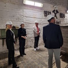 Vier Personen mit Schutzhelmen stehen in einem unterirdischen Tunnel mit weißen Backsteinwänden und Schienen am Boden und betrachten eine Wandmarkierung mit gekreuzten Hämmern und der Jahreszahl 1952
