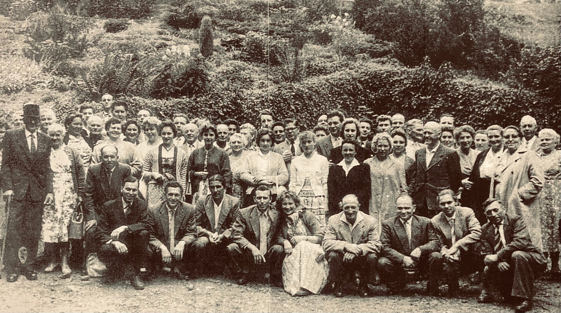 Historische Schwarz-Weiß-Gruppenaufnahme aus den 1950er-Jahren mit zahlreichen Frauen und Männern unterschiedlichen Alters. Die Personen stehen und knien in mehreren Reihen im Freien vor einer dicht bewachsenen Böschung