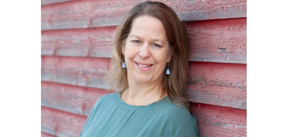 Portraits Clown Katharina Müller Lächelnde Frau mit blonden Haaren vor einer roten Holzwand.