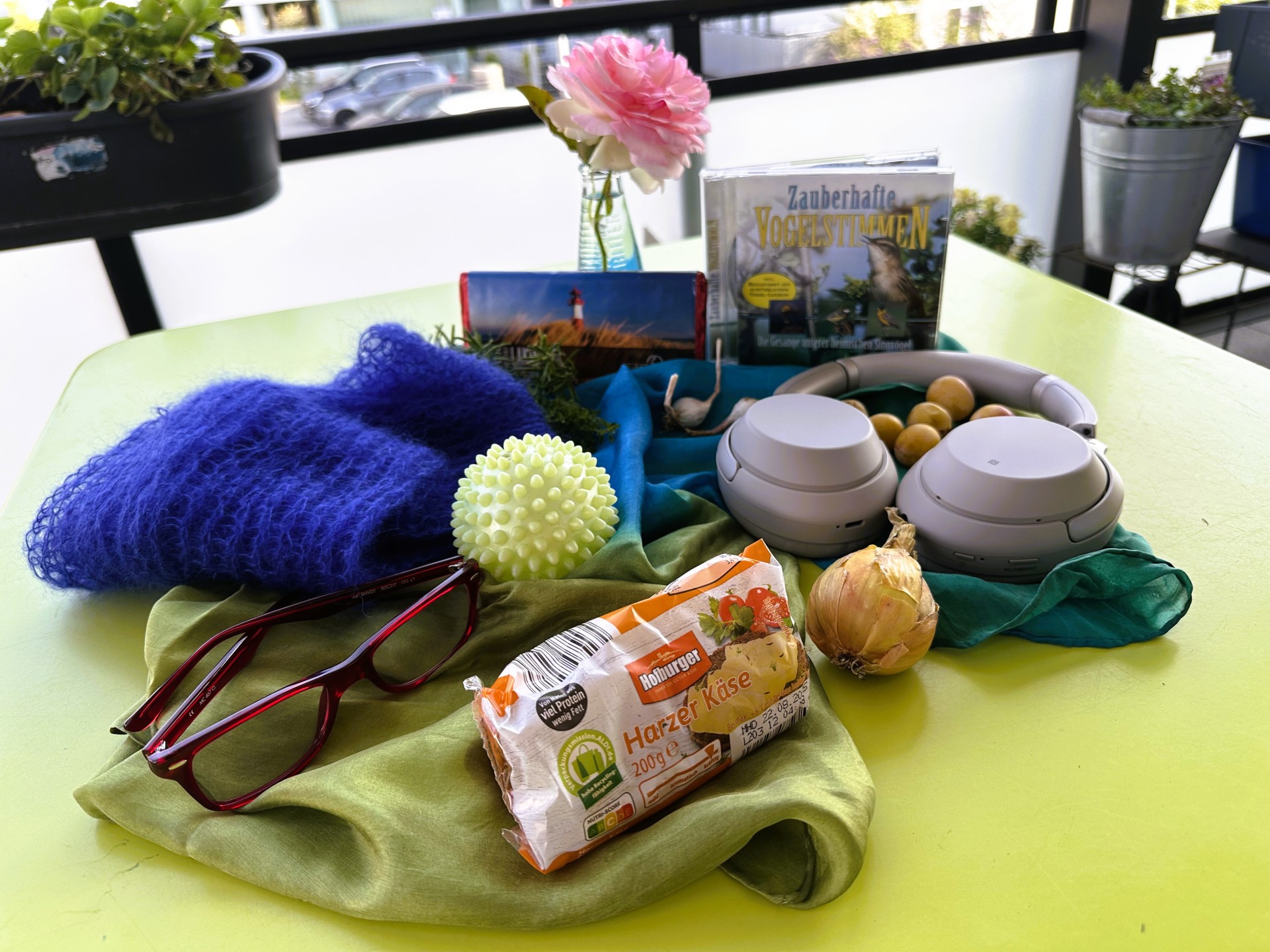 Die Blaue Stunde in der Petri-Villa Verschiedene Alltagsgegenstände – darunter Brille, Käse, Zwiebel, Kopfhörer und CDs – liegen auf einem gelbgrünen Tisch auf einem Balkon