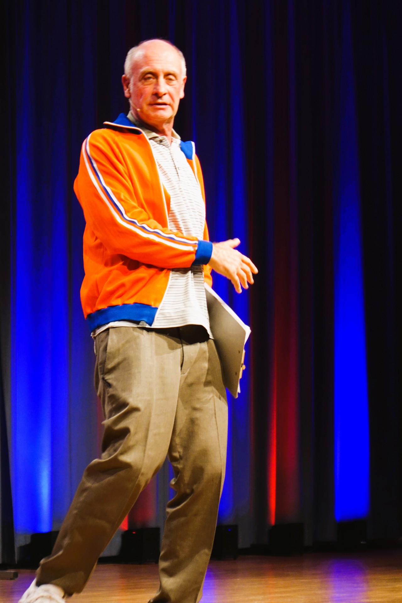 Alfons begeisterte in der Stadthalle in Ober-Ramstadt Ein Mann in orangefarbener Trainingsjacke steht auf einer Bühne und spricht mit einem Headset zum Publikum. In einer Hand hält er eine Mappe; der Bühnenvorhang im Hintergrund ist blau und rot beleuchtet.