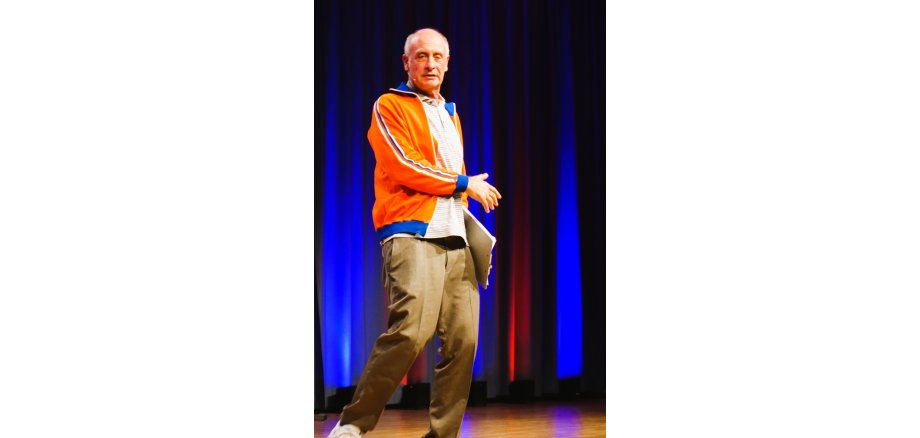 Alfons begeisterte in der Stadthalle in Ober-Ramstadt Ein Mann in orangefarbener Trainingsjacke steht auf einer Bühne und spricht mit einem Headset zum Publikum. In einer Hand hält er eine Mappe; der Bühnenvorhang im Hintergrund ist blau und rot beleuchtet.