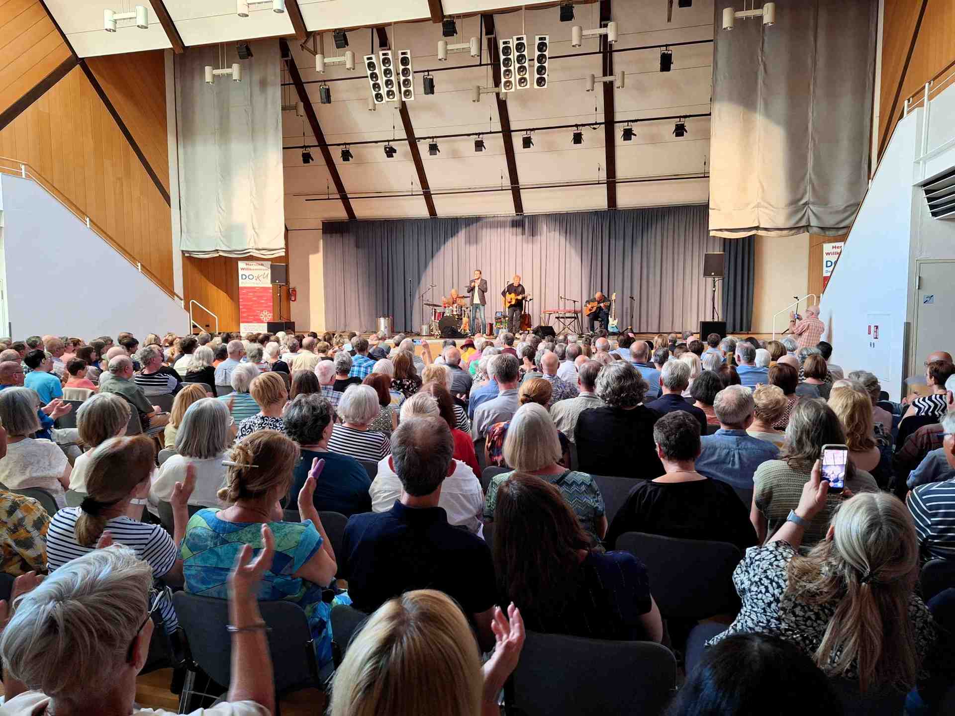 Auf einer Bühne spielt eine Musikgruppe vor einem voll besetzten Saal, während das Publikum aufmerksam zuhört und teilweise applaudiert