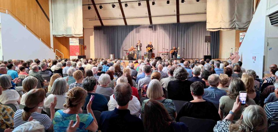 Auf einer Bühne spielt eine Musikgruppe vor einem voll besetzten Saal, während das Publikum aufmerksam zuhört und teilweise applaudiert