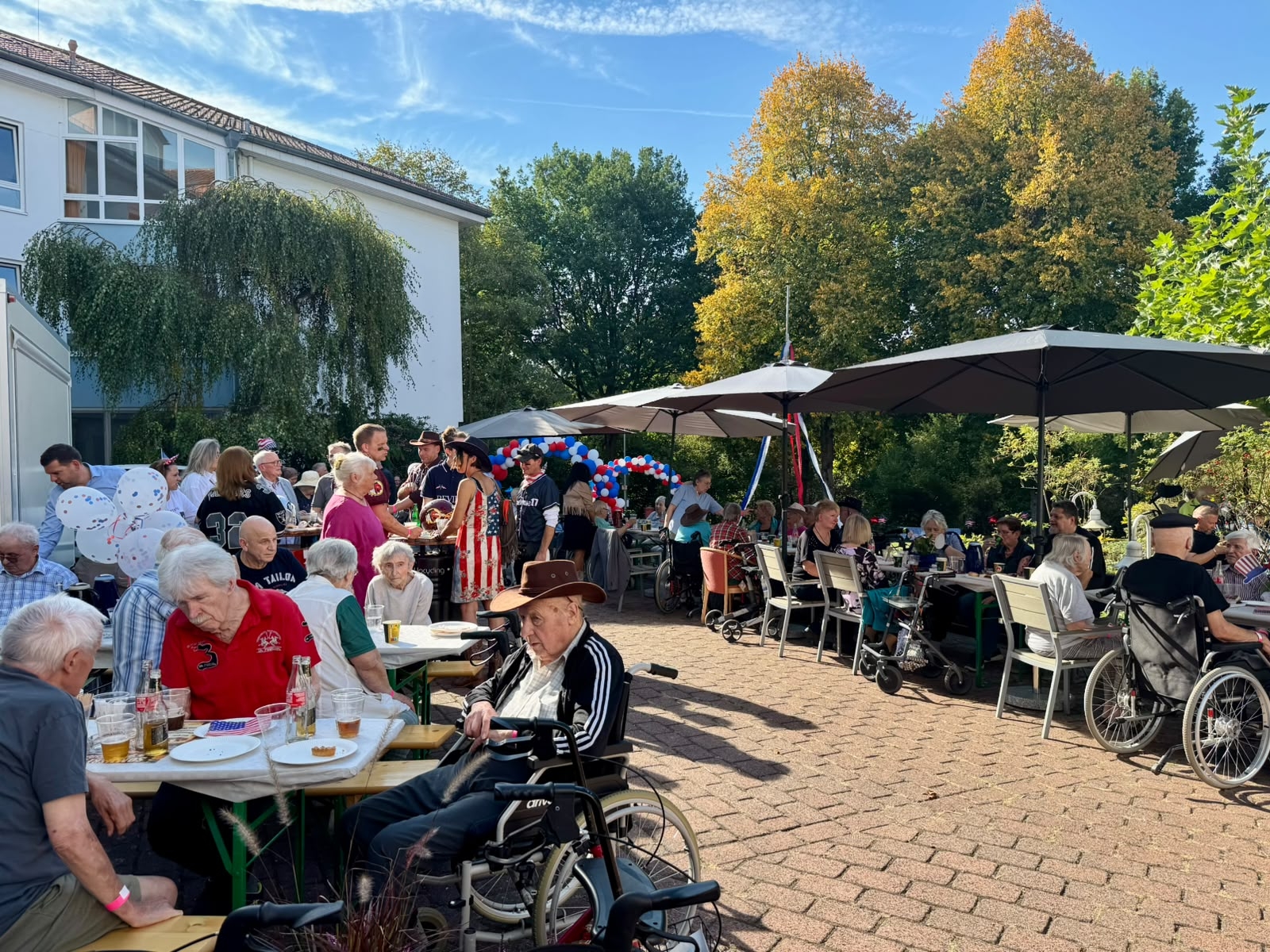 Die Bewohnerinnen und Bewohner des Heinrich-Gerold-Hauses genießen den Amerikanischen Flair. Zahlreiche Menschen sitzen und stehen bei einem Sommerfest im Freien an Tischen unter großen Sonnenschirmen. Der Bereich ist mit rot-weiß-blauen Luftballons und amerikanischen Flaggen dekoriert.