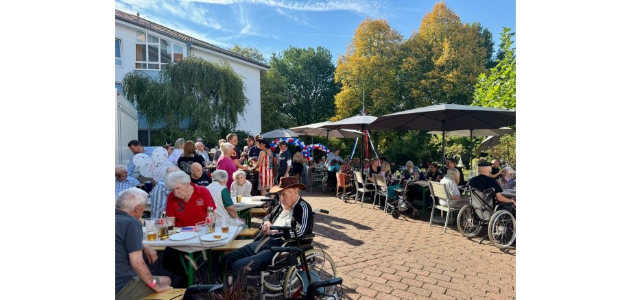 Die Bewohnerinnen und Bewohner des Heinrich-Gerold-Hauses genießen den Amerikanischen Flair. Zahlreiche Menschen sitzen und stehen bei einem Sommerfest im Freien an Tischen unter großen Sonnenschirmen. Der Bereich ist mit rot-weiß-blauen Luftballons und amerikanischen Flaggen dekoriert.