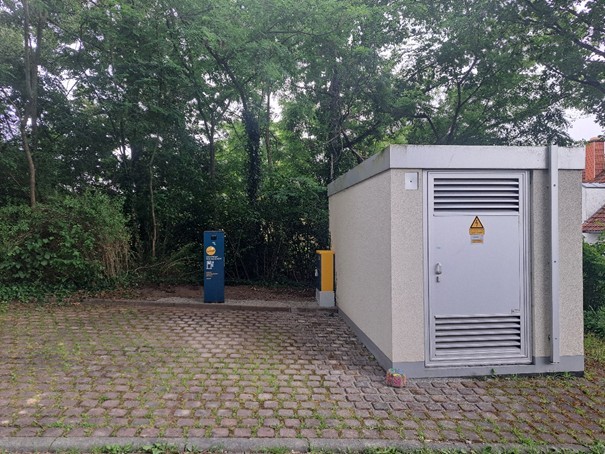 Im Wohngebiet Eiche Ost, steht jetzt eine neue E-Ladesäule Ein gepflasterter Platz vor einem kleinen Technikgebäude mit einer grauen Metalltür und Warnschild. Links daneben steht eine blau-gelbe Ladesäule für Elektrofahrzeuge.
