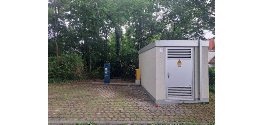 Ein gepflasterter Platz vor einem kleinen Technikgebäude mit einer grauen Metalltür und Warnschild. Links daneben steht eine blau-gelbe Ladesäule für Elektrofahrzeuge.