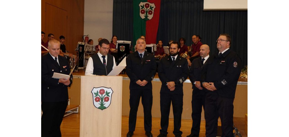 Bürgermeister Tobias Silbereis nimmt die Ehrungen vor. Sechs Männer in Feuerwehruniform stehen bei einer feierlichen Veranstaltung vor einer Bühne. Einer der Männer liest am Rednerpult aus einer Urkunde vor. Im Hintergrund sitzt ein Orchester mit Instrumenten und Notenständern.