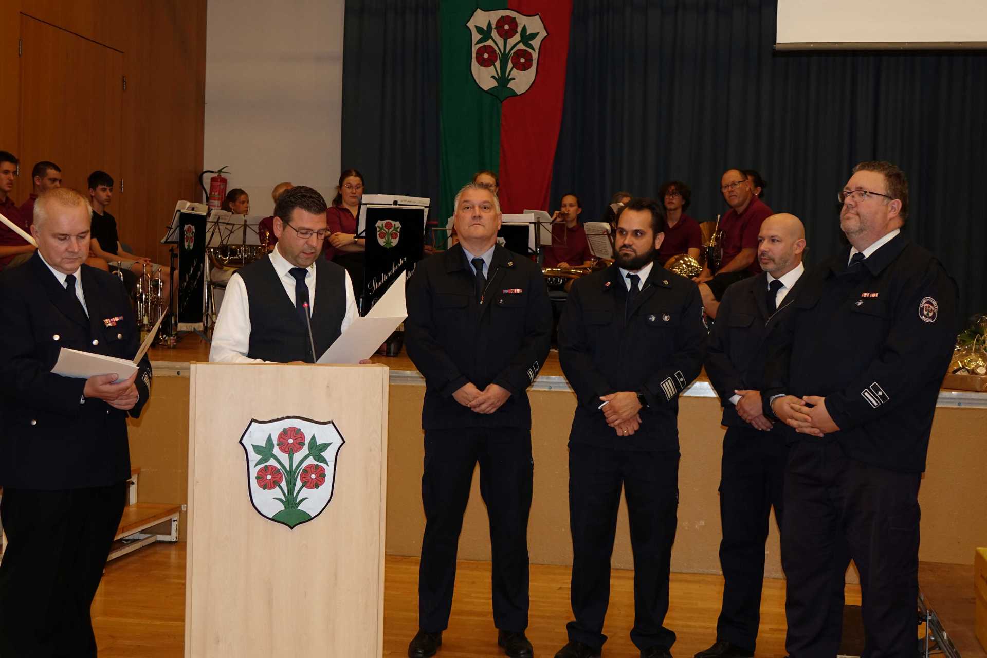 Sechs Männer in Feuerwehruniform stehen bei einer feierlichen Veranstaltung vor einer Bühne. Einer der Männer liest am Rednerpult aus einer Urkunde vor. Im Hintergrund sitzt ein Orchester mit Instrumenten und Notenständern.