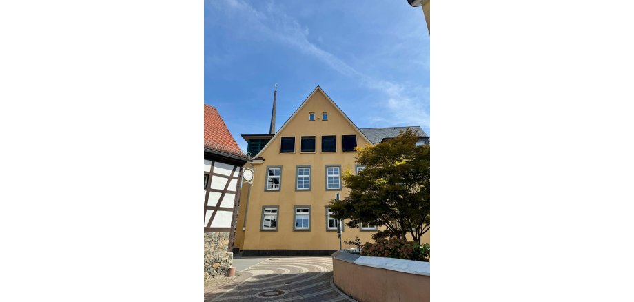 Die Außenfassade des Rathauses in Ober-Ramstadt Gelbe Gebäudefassade mit Giebeldach an einem gepflasterten Platz, eingerahmt von einem Fachwerkhaus und einem Baum.