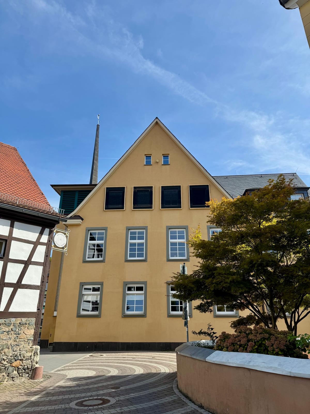 Gelbe Gebäudefassade mit Giebeldach an einem gepflasterten Platz, eingerahmt von einem Fachwerkhaus und einem Baum.