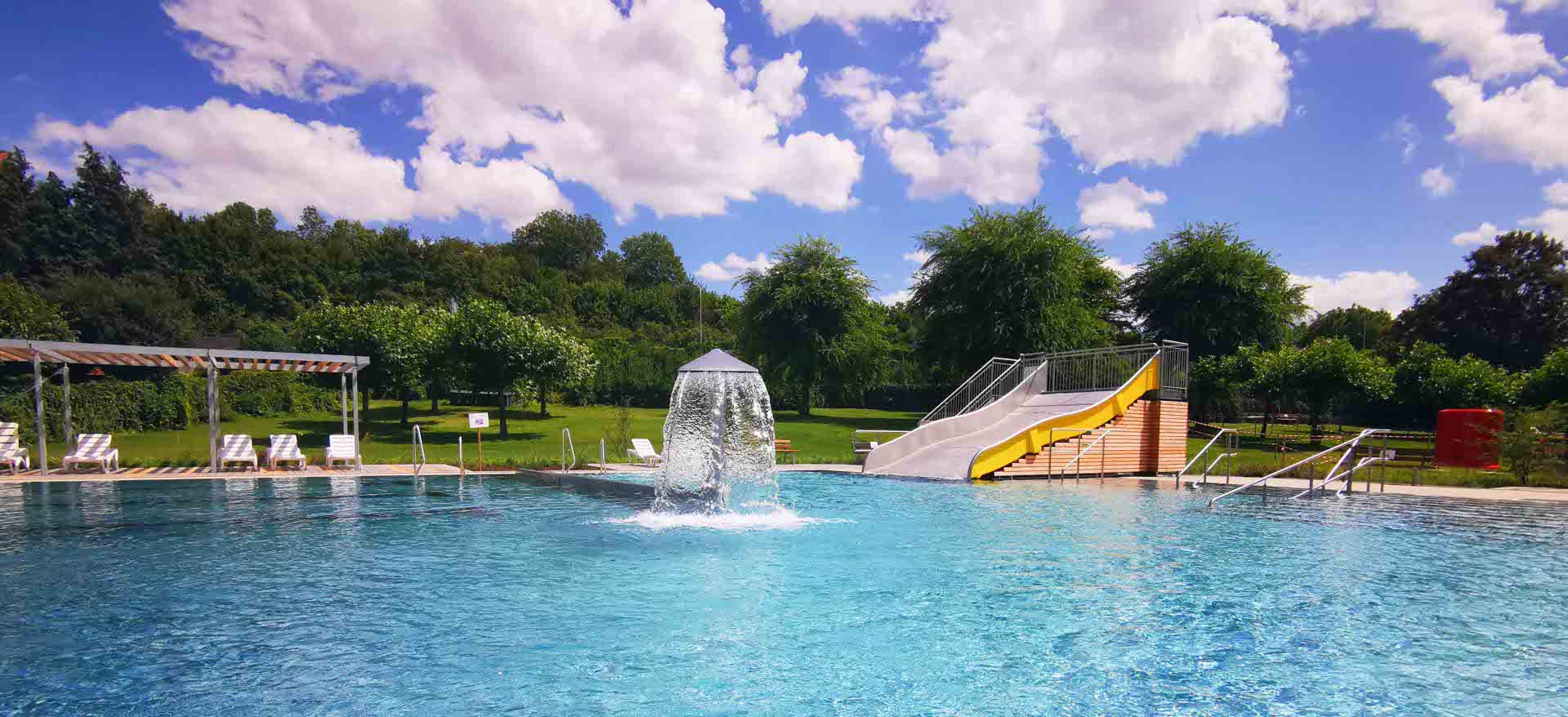 Das Bild zeigt das Freibad mit einer Rutsche, grüne Wiese und Sonnenliegen