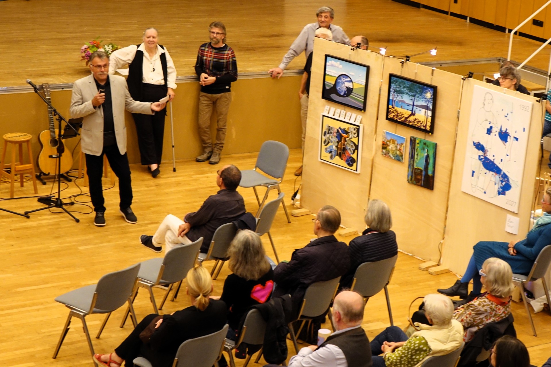 Magistratsmitglied Horst Becht eröffnet mit einer kleinen Ansprache die Herbstausstellung 2026 Ein Mann spricht vor sitzendem Publikum in einer Veranstaltungshalle. Neben ihm sind mehrere Kunstwerke auf Stellwänden ausgestellt; im Hintergrund stehen weitere Personen.