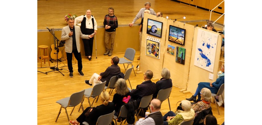 Magistratsmitglied Horst Becht eröffnet mit einer kleinen Ansprache die Herbstausstellung 2026 Ein Mann spricht vor sitzendem Publikum in einer Veranstaltungshalle. Neben ihm sind mehrere Kunstwerke auf Stellwänden ausgestellt; im Hintergrund stehen weitere Personen.