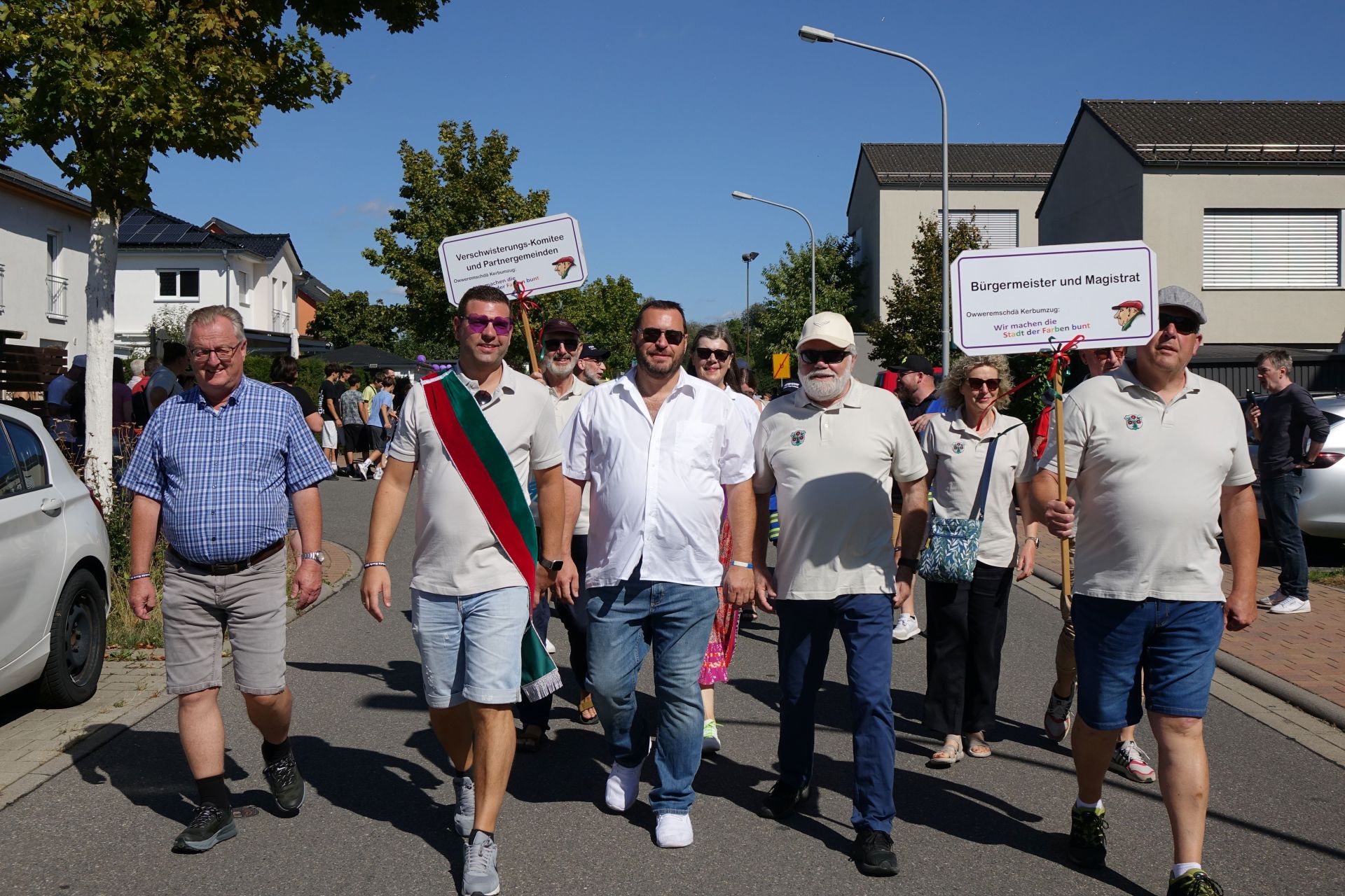 Mehrere Personen laufen bei einem Festumzug durch eine Wohnstraße. Zwei Teilnehmer tragen Schilder für das Verschwisterungs-Komitee sowie für Bürgermeister und Magistrat; im Hintergrund folgen weitere Menschen.