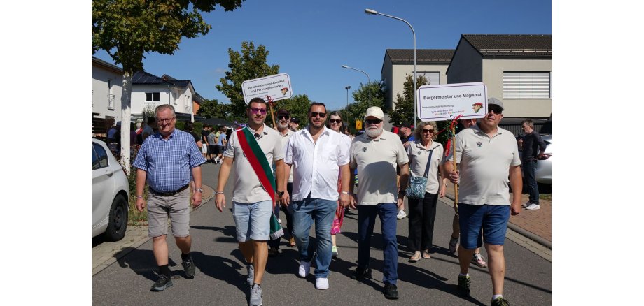 Bei sonnigem Wetter macht der Kerbumzug doch Spaß. Mehrere Personen laufen bei einem Festumzug durch eine Wohnstraße. Zwei Teilnehmer tragen Schilder für das Verschwisterungs-Komitee sowie für Bürgermeister und Magistrat; im Hintergrund folgen weitere Menschen.