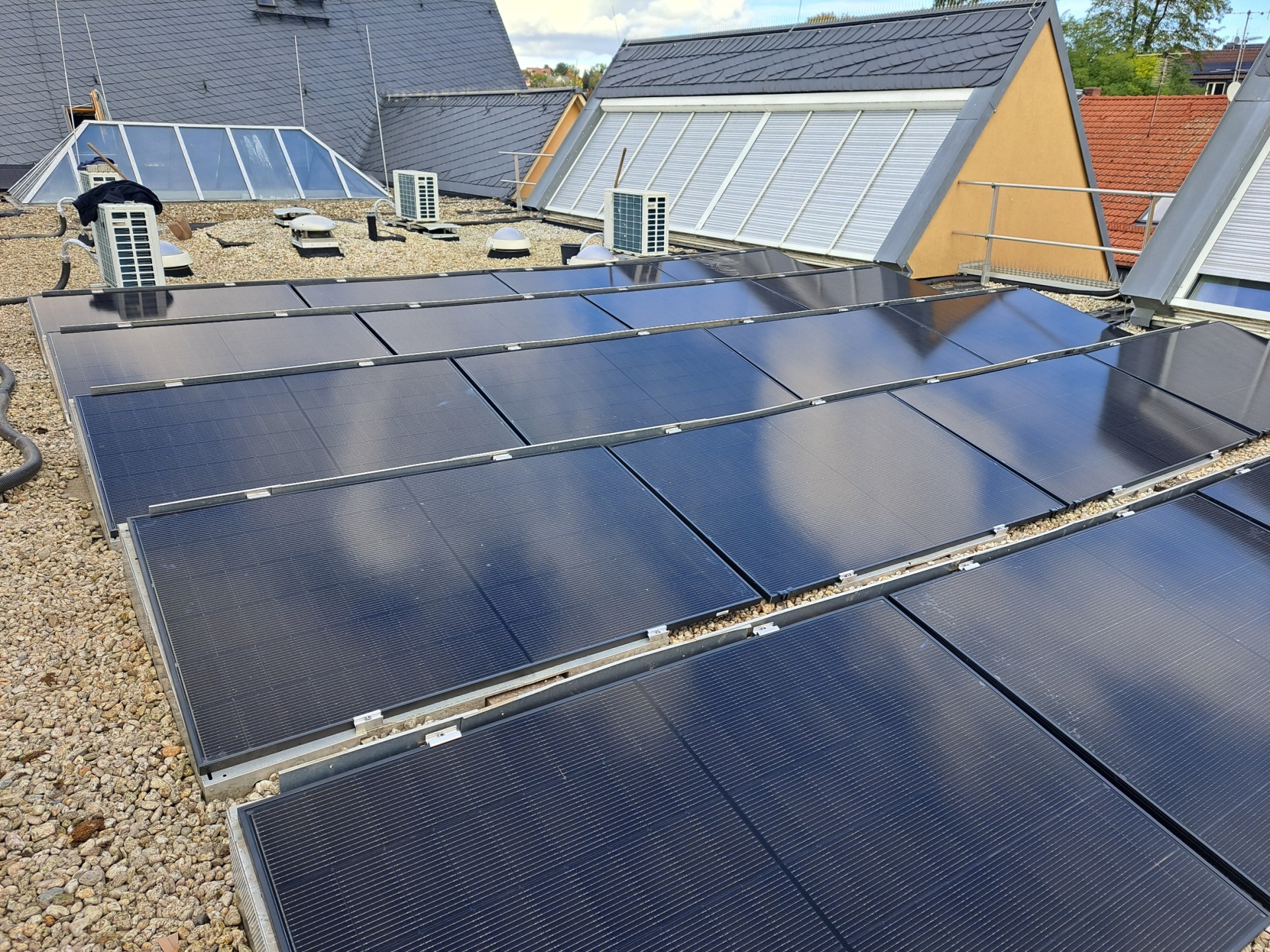 Mehrere Reihen dunkler Photovoltaikmodule auf einem flachen, mit Kies bedeckten Gebäudedach. Im Hintergrund sind Dachfenster, Lüftungsanlagen und angrenzende Gebäudedächer zu sehen.