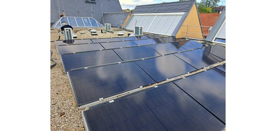 Die fertiggestellten PV-Module auf dem Rathausdacht Mehrere Reihen dunkler Photovoltaikmodule auf einem flachen, mit Kies bedeckten Gebäudedach. Im Hintergrund sind Dachfenster, Lüftungsanlagen und angrenzende Gebäudedächer zu sehen.