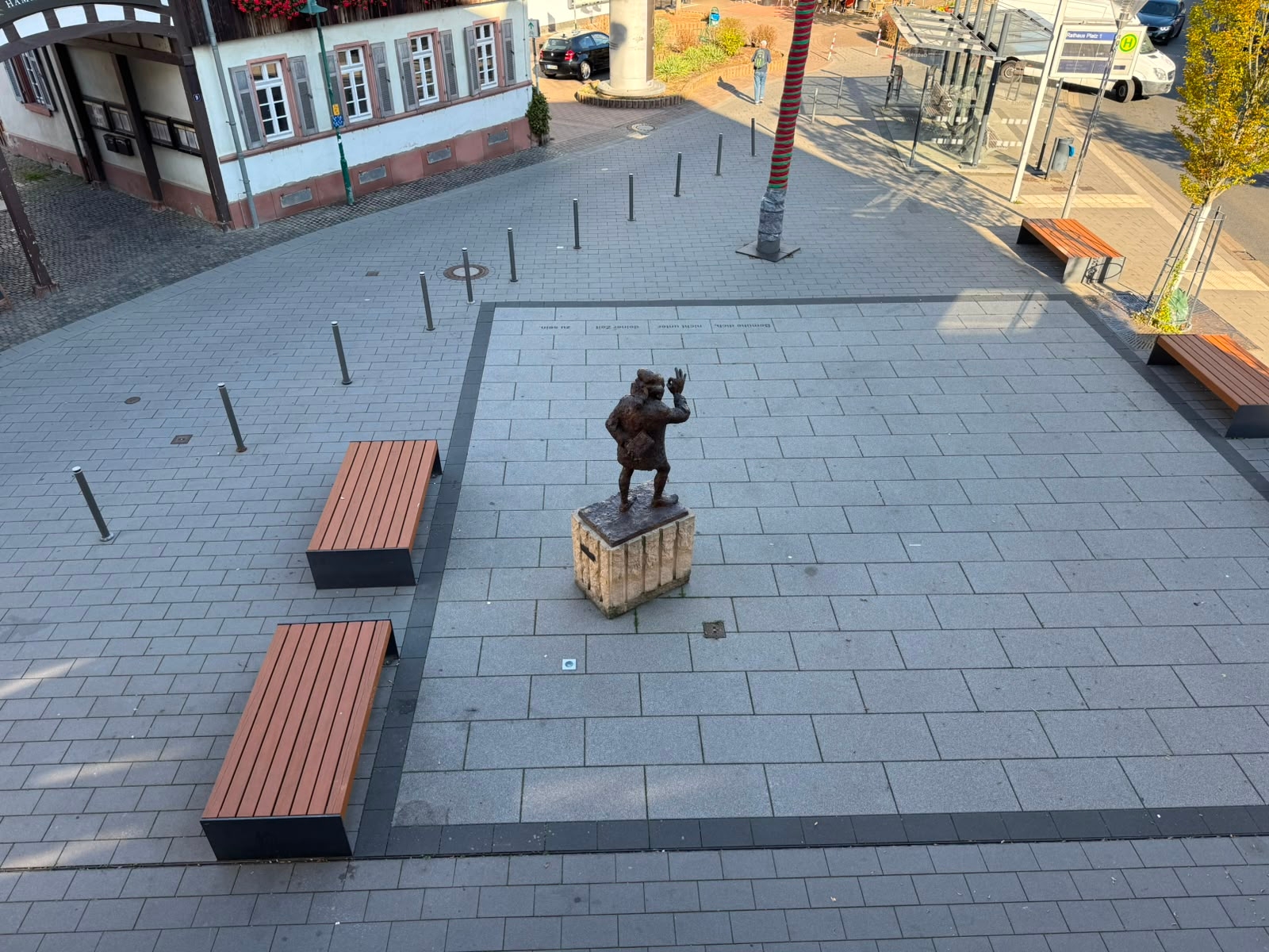 Der Rathausvorplatz in Ober-Ramstadt Blick von oben auf einen gepflasterten Rathausvorplatz mit einer Bronzefigur auf einem Steinsockel in der Mitte. Mehrere Holzbänke, Bäume und Poller säumen den Platz; im Hintergrund befinden sich eine Bushaltestelle und angrenzende Gebäude.