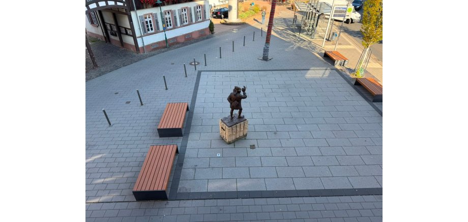 Der Rathausvorplatz in Ober-Ramstadt Blick von oben auf einen gepflasterten Rathausvorplatz mit einer Bronzefigur auf einem Steinsockel in der Mitte. Mehrere Holzbänke, Bäume und Poller säumen den Platz; im Hintergrund befinden sich eine Bushaltestelle und angrenzende Gebäude.