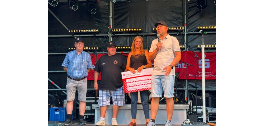 Feierliche Überreichung des Schecks an das Verschwisterungskomitee Vier Personen stehen auf einer Veranstaltungsbühne. Eine Frau hält einen großen Spendenscheck über 2.000 Euro, während der Mann neben ihr in ein Mikrofon spricht.