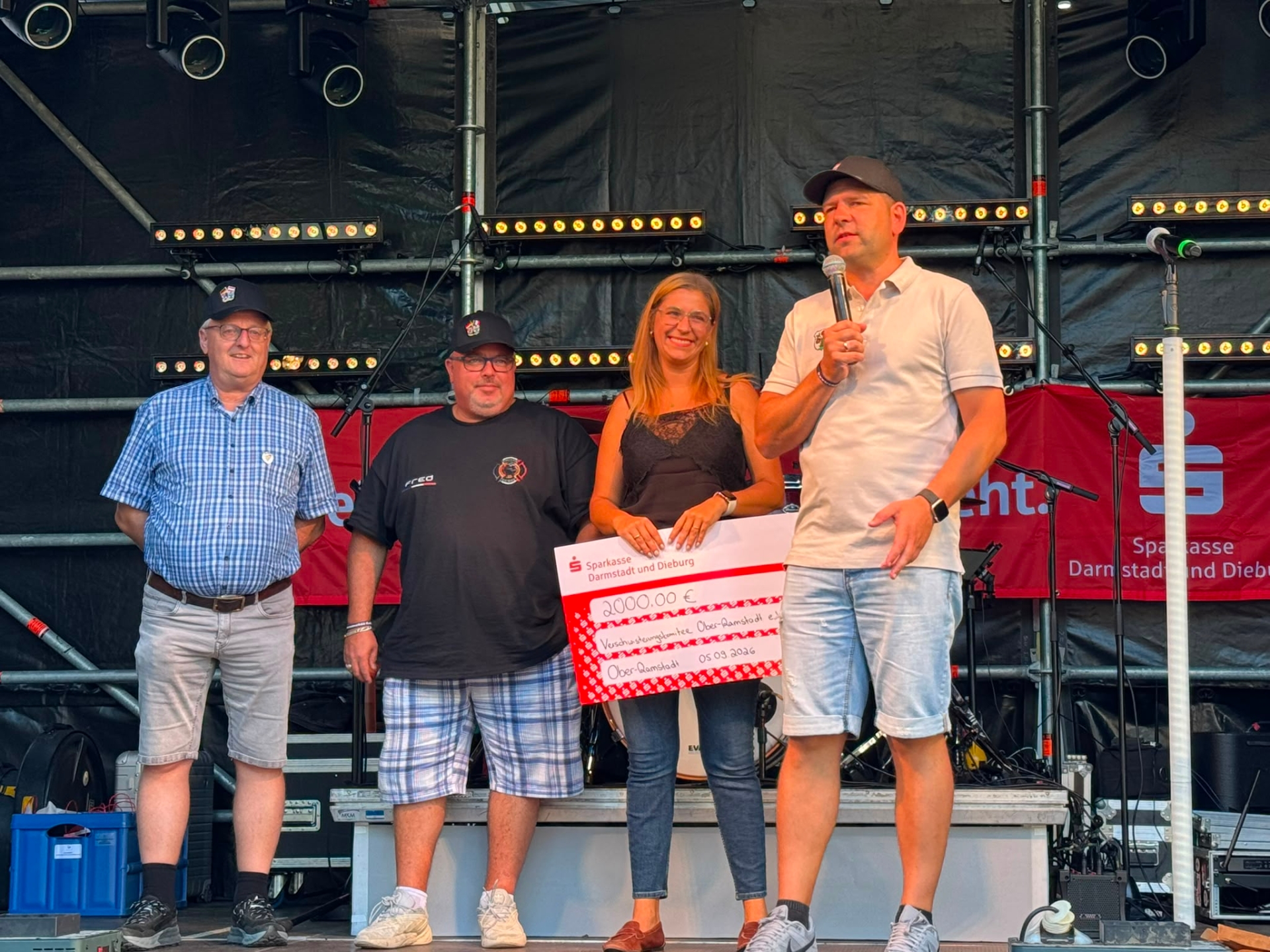 Vier Personen stehen auf einer Veranstaltungsbühne. Eine Frau hält einen großen Spendenscheck über 2.000 Euro, während der Mann neben ihr in ein Mikrofon spricht.