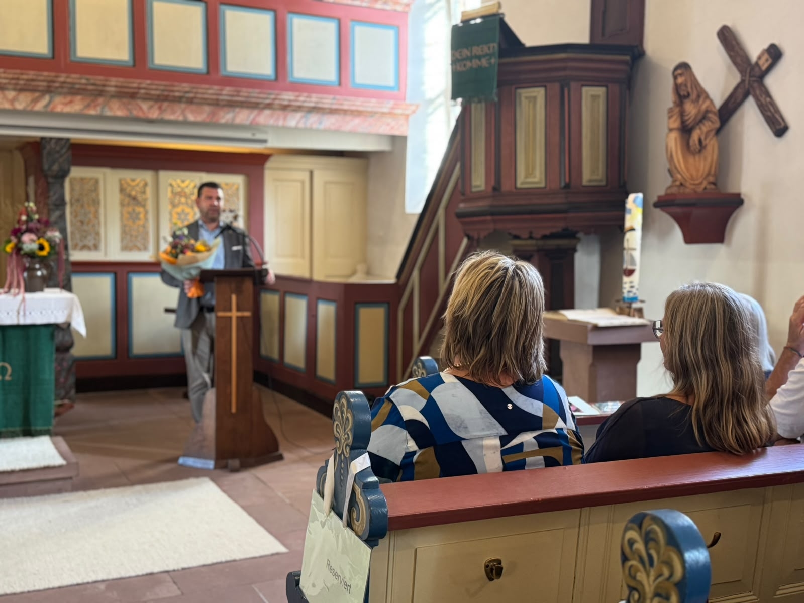 Ein Mann steht mit einem Blumenstrauß an einem Rednerpult in einer Kirche und spricht zu den Gästen. Im Vordergrund sitzen mehrere Personen in den Kirchenbänken.