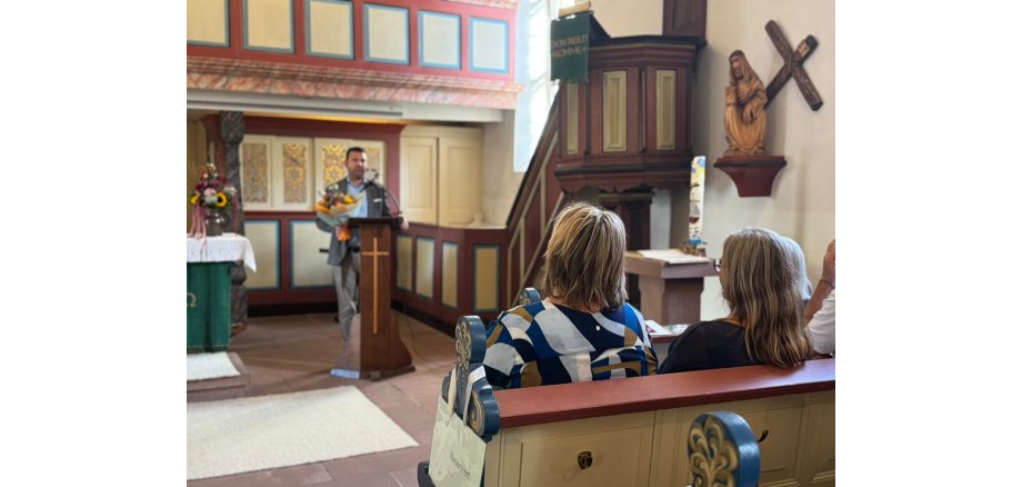Bürgermeister Tobias Silbereis verabschiedet Ute Lang und dankt ihr für ihr großes Engagement. Ein Mann steht mit einem Blumenstrauß an einem Rednerpult in einer Kirche und spricht zu den Gästen. Im Vordergrund sitzen mehrere Personen in den Kirchenbänken.