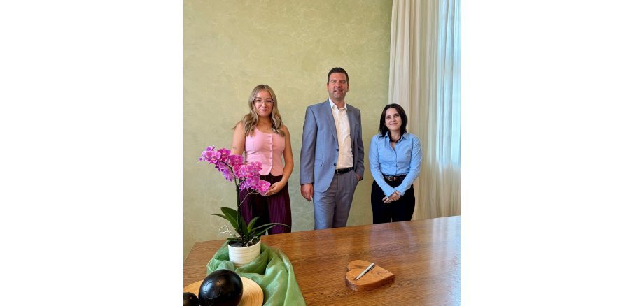 Tobias Silbereis zusammen mit den neuen Mitarbeiterinnen im Standesamt Drei Personen stehen nebeneinander in einem Standesamt hinter einem großen Holztisch. Im Vordergrund befindet sich eine pink blühende Orchidee; auf dem Tisch liegen außerdem ein Stift und eine herzförmige Holzablage.