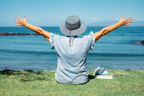 Back view of senior woman in hat sitting in meadow in front of sea looking at the horizon, relaxed senior lady enjoying free time on vacation or retirement